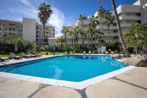 Piscina de un complejo turístico con tumbonas y palmeras, rodeada de edificios blancos de apartamentos de varias plantas bajo un cielo azul